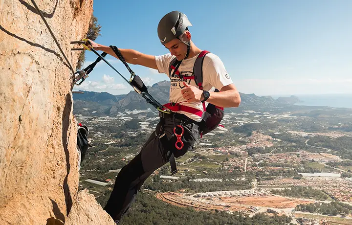 Deportista ascendiendo la vía ferrata del Ponoig en Alicante, con vistas panorámicas hacia Benidorm. Ruta vertical de 160 metros de altura con tramo final en descenso de rapel. Aventura y adrenalina en un entorno natural espectacular.
